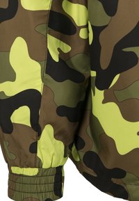 Kamouflagejacka med ett mönster i grönt, svart och gult, tillverkad av ett mjukt tyg med elastiska ärmslut för en åtsittande design.