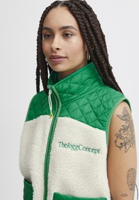 Chaleco acolchado verde con secciones de piel sintética crema, que presenta el logo "The/oggConcept" en bordado verde, bolsillo frontal y cuello alto.