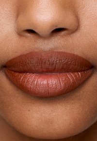 Matte brun leppestift påførte fullstendige, veldefinerte lepper med synlig tekstur og naturlige folder, mot en jevn hudbakgrunn.