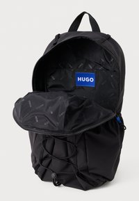 Juodas kuprinė su užtrauktuku, tekstūruota medžiaga ir mėlyna "HUGO" etikete viduje. Savybės - elastingos virvės, leidžiančios reguliuoti uždarymą.