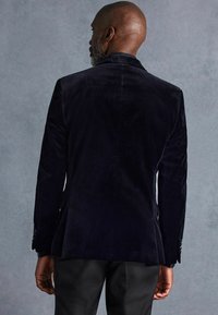 Blazer de terciopelo negro con un ajuste entallado, solapas puntiagudas y dos botones en los puños. Textura suave, adecuada para ocasiones formales.
