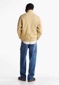 Felpa beige in pile con colletto alto, indossata sopra una camicia bianca, abbinata a pantaloni cargo in denim blu larghi. Scarpe marroni comode.