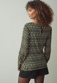 Blusa a maniche lunghe con un motivo a onde verde e crema, caratterizzata da un'apertura a occhio di bue sul retro e una texture liscia. Abbinata a pantaloni corti neri.