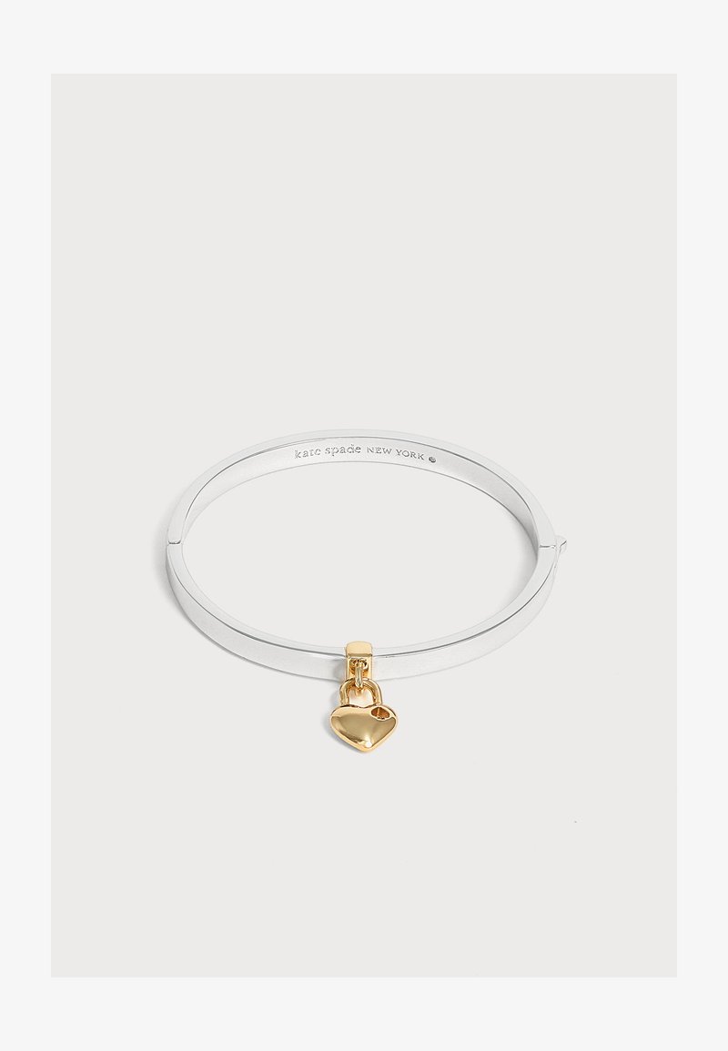 Bracelet jonc en argent avec un petit pendentif en forme de cadenas cœur doré suspendu dessus sur un fond blanc.