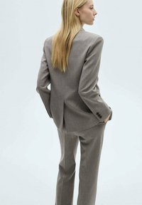Costume gris ajusté avec un blazer structuré et un pantalon à jambes droites. Tissu texturé, fente unique à l'arrière, détail de poignet à quatre boutons.