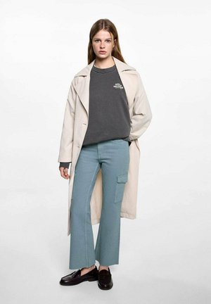 Béžový trenčkot přes šedou mikinu s logem, v kombinaci se světle modrými cargo kalhotami a černými mokasíny. Hladká látka, uvolněný styl.