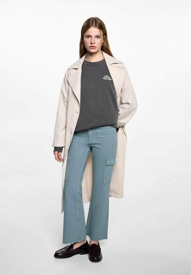 Béžový trench coat cez šedú mikinu s logom, v kombinácii s svetlomodrými cargo nohavicami a čiernymi mokasínami. Hladká tkanina, voľný strih.