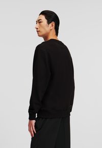 Felpa nera con scollo rotondo, polsini e orlo a coste. Presenta una texture liscia e una vestibilità comoda, indossata con pantaloni neri.