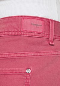 Jean en denim rose avec une étiquette indiquant "Pepe Jeans". Comprend une poche arrière avec rivet en métal et un tissu texturé.