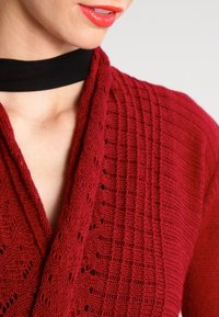Rote gestrickte Strickjacke mit einem strukturierten Design und offener Vorderseite, mit ausgearbeiteten Mustern im Stoff und einem schwarzen Choker-Ausschnitt.