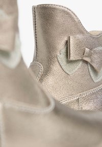 Bottines en cuir doré métallique avec une texture lisse, mettant en avant un accent de nœud proéminent et des détails de couleur claire contrastants.