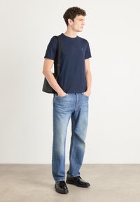Tricou navy cu guler rotund, blugi drept de culoare bleu deschis și pantofi negri. Modelul ține o geantă tote neagră, stând pe un fundal neutru.