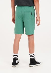 Groene katoenen shorts met een achterzak, elastische tailleband en een relaxte pasvorm, gecombineerd met zwarte hoge sneakers en gestreepte sokken.