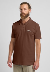 Polo marrone con colletto, maniche corte e logo sul petto a sinistra. Il tessuto appare liscio con una trama e una vestibilità uniformi.