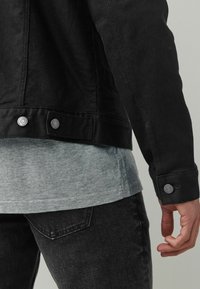 Chaqueta de mezclilla negra con botones de metal, llevada sobre una camiseta gris. Incluye un tejido texturizado y un diseño ajustado, mostrada desde la parte posterior.