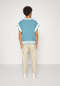Polo a maniche corte in maglia blu con rifiniture bianche; presenta una fascia a coste a contrasto con la scritta "Emporio Armani." Pantaloni beige.