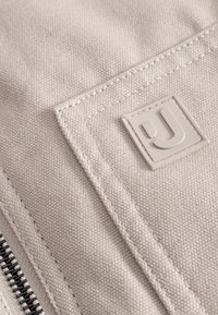 Beżowa tkanina o teksturowanej powierzchni, z prostokątną jasnoszara łatą z logo oraz ciemnym zamkiem błyskawicznym wzdłuż krawędzi.