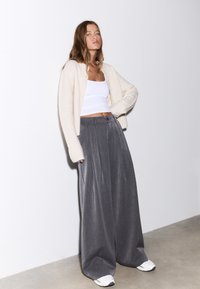 Pantalon gris à jambes larges associé à un cardigan crème et un débardeur blanc. Le pantalon présente des plis ; la tenue est complétée par des baskets blanches.
