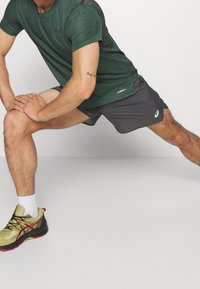 Green perforated athletic shirt, black checkered shorts, and olive running shoes with red accents and black sole; engaged in a stretching pose.