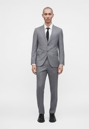 Mężczyzna ubrany w szary garnitur w cienkie prążki z dopasowanymi spodniami, białą koszulę, czarny krawat i czarne eleganckie buty, stojący na białym tle.
