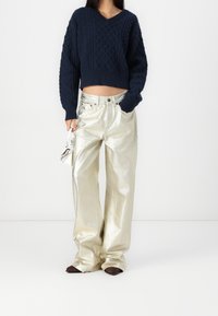 Granatowy sweter dzianinowy z wzorem w kabel, w połączeniu z wysokimi, szerokimi spodniami metalicznymi w kolorze złotym. W ręku elegancka biała torebka.