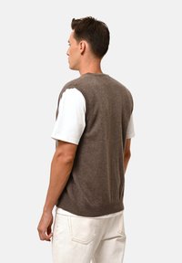 Brun ullväst med rund hals, ärmlös design och en slät textur, bärs över en vit t-shirt och matchas med ljusa byxor.