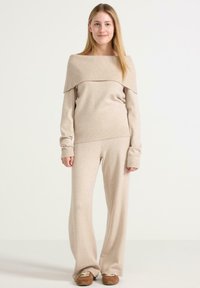 Junge Frau steht in einem beigen, schulterfreien Pullover, passenden weiten Hosen und braunen Freizeitschuhen vor einem weißen Hintergrund.