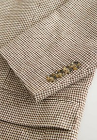 Rukáv hnedo-bieleho blejzra s kockovaným vzorom, so štyrmi gombíkmi z lišajníkového vzoru a s klapkovou vreckom na svetlom pozadí.