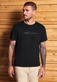 Muž s krátkými vlasy a tetováním na obou pažích nosí černé tričko "Crafted Goods" a béžové kalhoty, stojící proti pozadí ze dřevěných panelů.