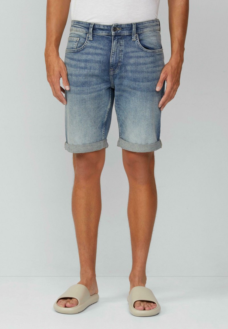 Shorts en denim bleu clair avec une finition délavée, ourlets repliés, design traditionnel à cinq poches et texture usée. Associés à des sandales à glissière beige.