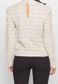 Beige textured top with perforated pattern, long sleeves, and a keyhole opening at the back. Fitted design with slight puffed shoulders.