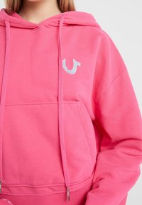 Sudadera rosa con un bolsillo frontal, capucha con cordón y acentuación de logotipo metálico. Tejido suave con una textura lisa, con un ajuste casual.