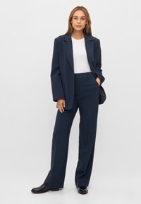 Marineblauwe op maat gemaakte kostuum met een dubbelbreasted blazer met brede revers, bijpassende hoge taillebroek en een witte aansluitende top eronder.