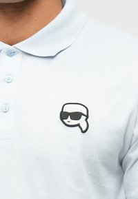 Polo bleu clair avec un petit patch noir et blanc d’un visage portant des lunettes de soleil sur la zone de la poitrine.
