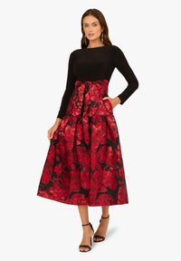 Schwarzes Langarmoberteil kombiniert mit einem rot floral gemusterten Rock, der eine Schleifendetail an der Taille aufweist, aus fließendem Stoff und begleitet von Sandalen mit Knöchelriemen.