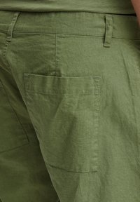 Žali lininiai krovini�ų stiliaus šortai su tekstūruota paviršiaus struktūra, turintys plokščią užpakalinę kišenę ir laisvą siluetą su matomomis siuvimo detalėmis.