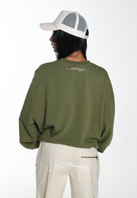 Oliwkowy, oversize'owy sweatshirt z okrągłym dekoltem i białym logo na plecach, zestawiony z beżowymi spodniami cargo z zasuwaną kieszenią.