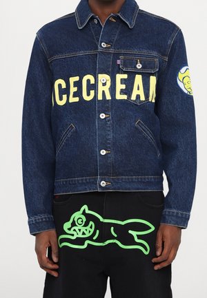 Person iført en mørk denimjakke med gul "ICECREAM" tekst og en stikl-logo-patch, kombineret med sorte bukser med en grøn tegneseriehund grafik.