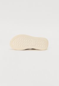 Semelle de chaussure en caoutchouc crème avec un motif de grip texturé, présentant des carrés en relief et des ondulations le long des bords. Marque visible sur la semelle.