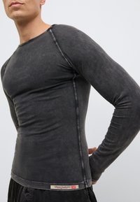 Čierna košeľa s dlhými rukávmi z mäkkej tkaniny so vzhľadom opotrebenia, s kontrastným prešívaním a malou štítkovou značkou pri leme.