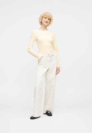 Νέα γυναίκα με κοντά σγουρά ξανθά μαλλιά, που φορά μπλούζα μακρυμάνικη σε απαλό κίτρινο χρώμα, λευκά παντελόνια φαρδιά με μικρά σχέδια και μαύρα φλατ παπούτσια.