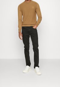 Brun stickad tröja med ruggad struktur, svarta jeans och vita sneakers. Tröjan har en liten logotyppatch på bröstet.