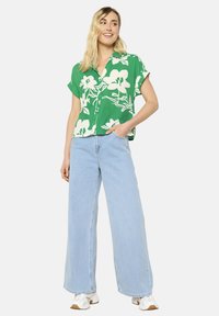 Camicia verde a maniche corte con motivi bianchi, abbinata a jeans a gamba larga blu chiaro e sneakers bianche. Il modello sta in posa in modo disinvolto con le mani in tasca.