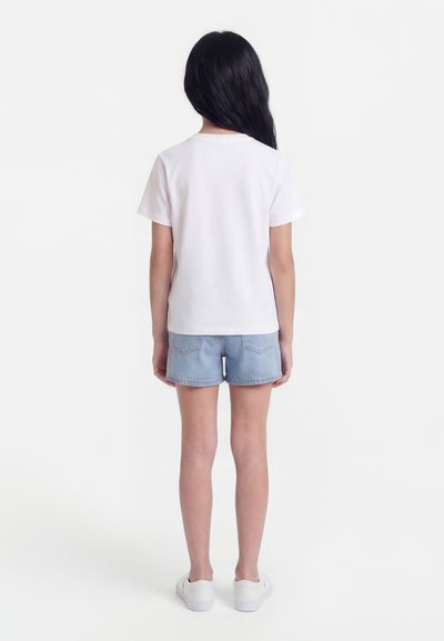 Child with long black hair wearing a white short-sleeve t-shirt, light blue denim shorts, and white sneakers, standing facing away.