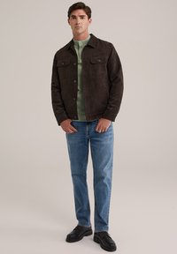 Sötétbarna velúr dzseki, két mellzsebbel, világos zöld felső fölött viselve, kék farmerrel és fekete cipővel párosítva. Semleges háttér.