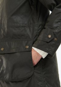 Giacca in tessuto cerato verde scuro con una tasca a toppa, polsini con bottone e foderatura interna in maglia bianca. Branding discreto visibile.