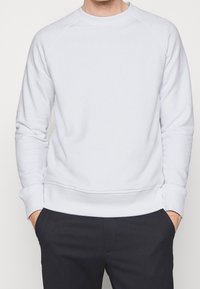 Sudadera blanca con cuello redondo y mangas raglán, con dobladillo y puños acanalados. Textura suave, combinada con pantalones oscuros.