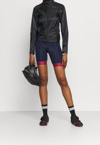 Czarna kurtka typu windbreaker z zamkiem, granatowe szorty rowerowe z czerwonymi akcentami, czarne buty rowerowe, trzyma czarny kask.