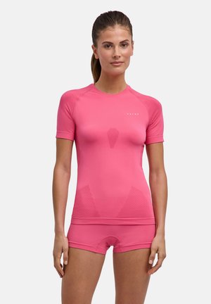 Vrouw draagt een aansluitend roze sporttop met korte mouwen en bijpassende shorts, staand met handen ontspannen langs de zij tegen een effen achtergrond.