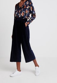 Mörkblå kordflamsbyxor med vida ben, tillsammans med en blus i flerfärgat djurmönster med V-ringning. Vita sneakers avslutar looken.